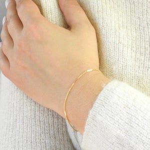 May include: A delicate gold bracelet with a fine chain design. The bracelet is worn on a wrist, and the gold color contrasts with the light-colored sweater. The bracelet appears to be a simple, elegant piece of jewelry.