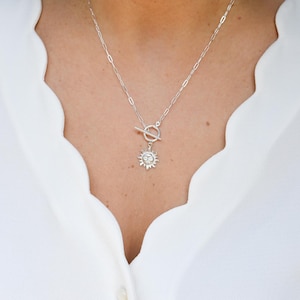 Op de afbeelding: Een zilveren ketting met een zonnehanger. De ketting heeft een tuimelsluiting en een fijne ketting. De zonnehanger heeft een gedetailleerd ontwerp met stralen en een centraal cirkelvormig element. De ketting wordt gedragen over een witte blouse.