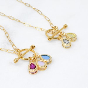 May include: Two gold-colored necklaces with a toggle clasp and teardrop-shaped pendants. Each pendant features three colorful gemstones in shades of pink, blue, and yellow. The necklaces are displayed on a white surface.