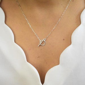 May include: A silver necklace with a toggle clasp closure. The necklace features a delicate chain and a circular pendant with a bar closure. The jewelry is worn against a white blouse.