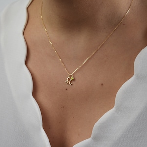 May include: A gold-colored necklace featuring a Leo zodiac symbol pendant and a small green gemstone charm. The delicate chain is visible against a white top, showcasing the jewelry's design and color.