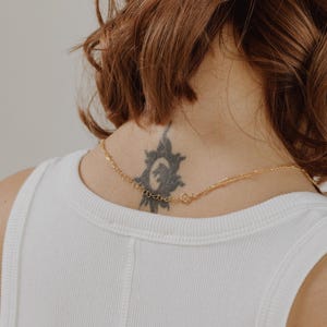 May include: A gold chain necklace with a delicate design. The necklace is worn by a person with a tattoo of a sun on their neck.