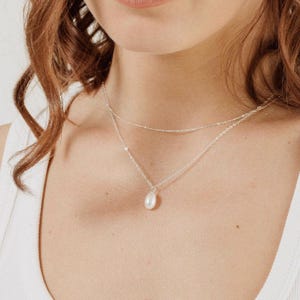 Può includere: Una collana a catena d'argento con un ciondolo a perla singola. La collana è sovrapposta a una seconda catena più sottile.