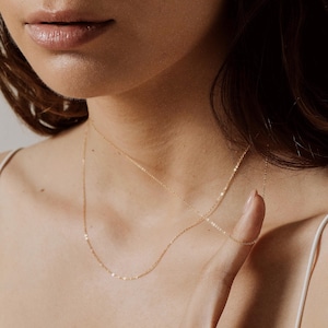 Peut inclure: Un collier fin en chaîne dorée avec un design simple. Le collier est superposé à une deuxième chaîne plus longue.