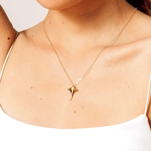 Könnte beinhalten: Eine goldene Halskette mit einem zarten, stilisierten Rochen-Anhänger. Die Kette wird von einer Person mit einem weißen Tanktop getragen.