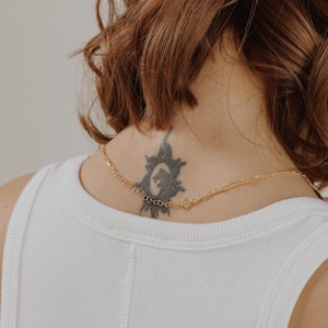 May include: A gold chain necklace with a delicate design. The necklace is worn by a person with a tattoo of a sun on their neck.