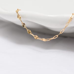 May include: A delicate gold necklace with a unique design. The necklace features a combination of small, round, textured elements and elongated, rectangular links. The jewelry is displayed on a white surface, highlighting its color and design.