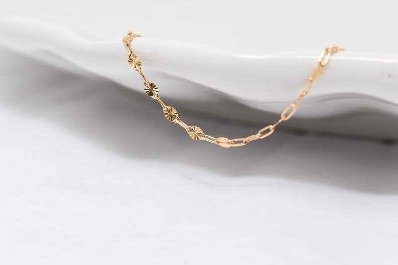 May include: A delicate gold necklace with a unique design. The necklace features a combination of small, round, textured elements and elongated, rectangular links. The jewelry is displayed on a white surface, highlighting its color and design.