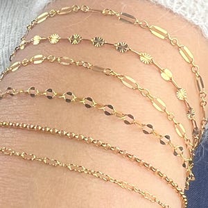 May include: Close-up of several delicate gold bracelets. The bracelets feature various chain styles, including oval links, small circles, and floral-shaped accents. The jewelry is displayed on a person's arm, showcasing the different designs and textures.