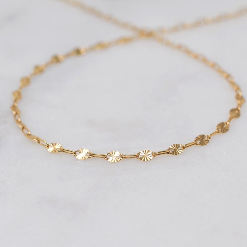 May include: A delicate gold necklace featuring a chain with small, circular, starburst-like charms. The necklace is displayed on a white surface, showcasing its intricate design and warm, golden hue. This piece is ideal for adding a touch of elegance.