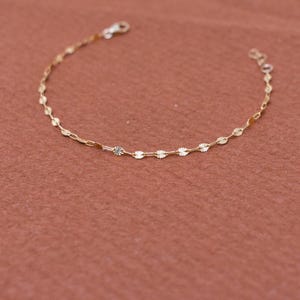 May include: Delicate gold bracelet featuring a repeating pattern of small, circular charms interspersed with oval links. The bracelet has a lobster clasp closure and is displayed against a warm, reddish-brown background. A simple and elegant piece of jewelry.