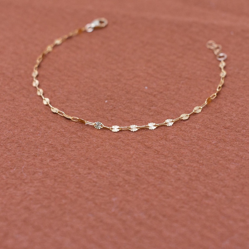 May include: Delicate gold bracelet featuring a repeating pattern of small, circular charms interspersed with oval links. The bracelet has a lobster clasp closure and is displayed against a warm, reddish-brown background. A simple and elegant piece of jewelry.