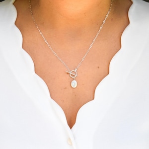 Op de afbeelding: Een zilveren ketting met een tuimelsluiting en een peervormige, iriserende hanger. De ketting wordt gedragen tegen een witte blouse met een geschulpte halslijn. De fijne ketting en de hanger creëren een eenvoudige, elegante look. De hanger is ongeveer 1,5 cm lang.