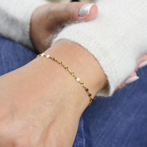 May include: A delicate gold bracelet featuring small, circular and petal-shaped charms. The bracelet is worn on a wrist, with a white sweater and blue jeans in the background. The bracelet is a simple, elegant piece of jewelry.