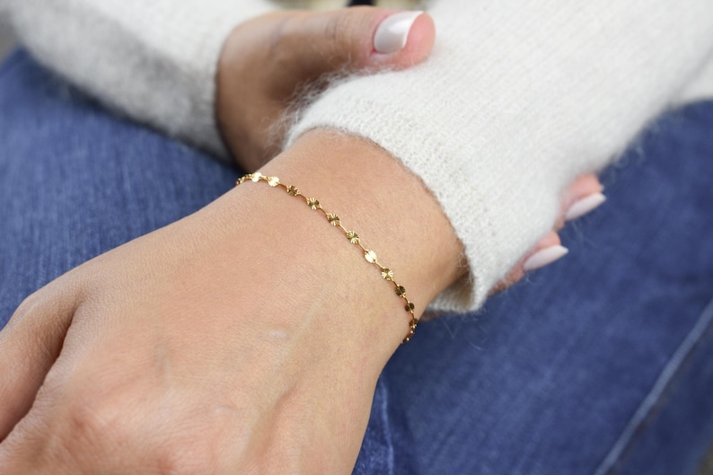 May include: A delicate gold bracelet featuring small, circular and petal-shaped charms. The bracelet is worn on a wrist, with a white sweater and blue jeans in the background. The bracelet is a simple, elegant piece of jewelry.