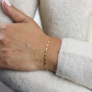 May include: A delicate gold bracelet with a repeating leaf-shaped design. The bracelet is worn on a wrist, and the background is a white sweater.