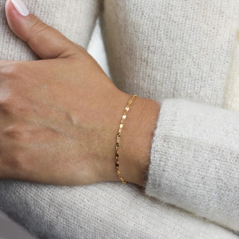 May include: A delicate gold bracelet with a repeating leaf-shaped design. The bracelet is worn on a wrist, and the background is a white sweater.