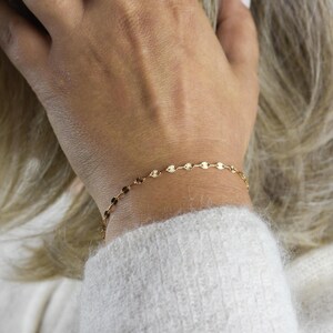 May include: Delicate gold bracelet featuring a series of small, circular charms. The bracelet is worn on a wrist, with a soft, white sweater in the background. The charms have a subtle, textured design, catching the light and adding a touch of elegance.