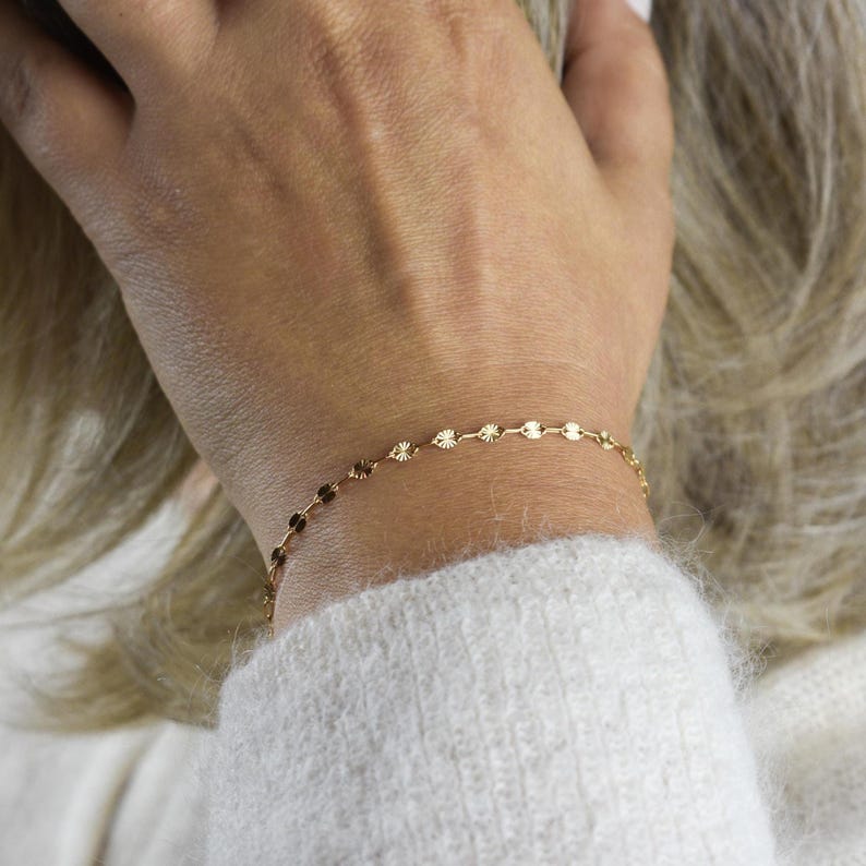 May include: Delicate gold bracelet featuring a series of small, circular charms. The bracelet is worn on a wrist, with a soft, white sweater in the background. The charms have a subtle, textured design, catching the light and adding a touch of elegance.
