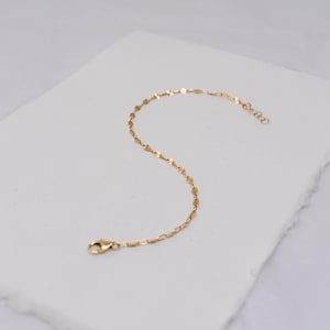 May include: A delicate gold-colored bracelet with a repeating pattern of small, flat, irregular shapes. The bracelet has a lobster clasp closure and a small chain for adjusting the length. The jewelry is displayed on a white surface.