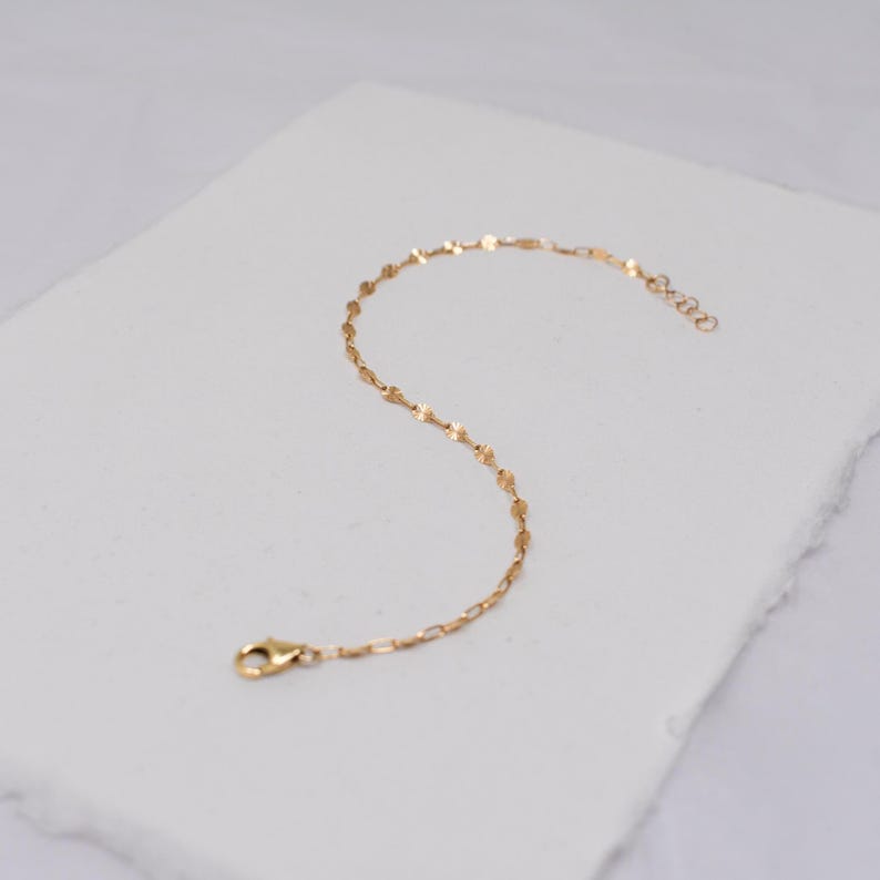 May include: A delicate gold-colored bracelet with a repeating pattern of small, flat, irregular shapes. The bracelet has a lobster clasp closure and a small chain for adjusting the length. The jewelry is displayed on a white surface.