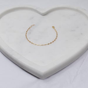 May include: Delicate gold bracelet featuring small, flat, and round charms. The bracelet is displayed on a white, heart-shaped marble tray. The jewelry is simple and elegant, suitable for everyday wear.