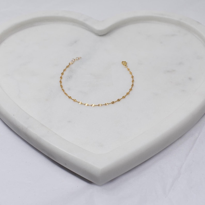 May include: Delicate gold bracelet featuring small, flat, and round charms. The bracelet is displayed on a white, heart-shaped marble tray. The jewelry is simple and elegant, suitable for everyday wear.