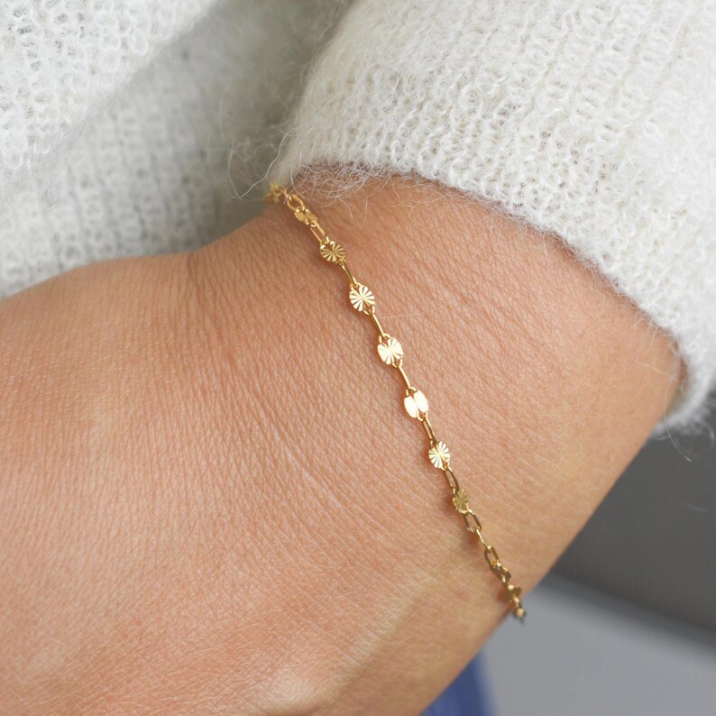 May include: A delicate gold bracelet featuring a chain of small, circular and starburst-shaped links. The bracelet is worn on a wrist, with a soft, white sweater in the background. The bracelet is a simple, elegant piece of jewelry.