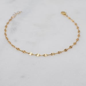 May include: Delicate gold-colored bracelet with a series of small, round, textured charms. The bracelet is made of thin chain links and has a lobster clasp closure. The jewelry is displayed on a white marble surface.