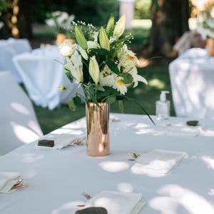 May include: A cylindrical copper vase holds a bouquet of white lilies, green foliage, and small white filler flowers. The vase sits on a white tablecloth set with white napkins, small grey stones, and metallic cutlery at each place setting.