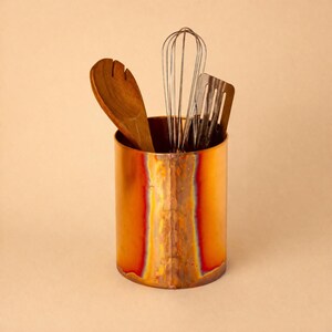 May include: A cylindrical copper utensil holder with a vertical heat-treated seam displaying iridescent orange, red, and purple tones. Inside the holder are a wooden salad server, a metal wire whisk, and a dark metal slotted spatula.