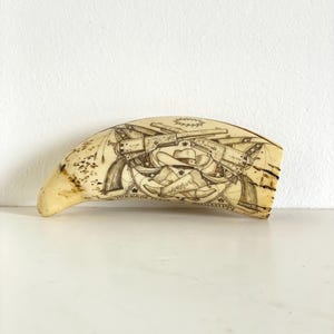 Puede incluir: Un colmillo tallado, de color blanquecino, con un grabado detallado. La obra de arte presenta un sombrero de vaquero, pistolas cruzadas, estrellas y texto. El colmillo tiene una forma curva y un aspecto liso y pulido.