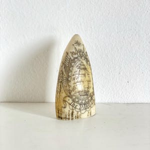 Könnte beinhalten: Ein cremefarbenes, geschnitztes Objekt mit spitzer Spitze. Das Objekt zeigt eine detaillierte Gravur eines Segelschiffs, Flaggen und die Worte "The Dakota". Das Objekt wird auf einer weißen Oberfläche präsentiert.