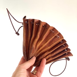 May include: A brown leather pouch with a drawstring closure. The pouch is fan-shaped and has a textured surface. The drawstring is a thin, dark brown cord. The pouch is held in a hand against a white background.