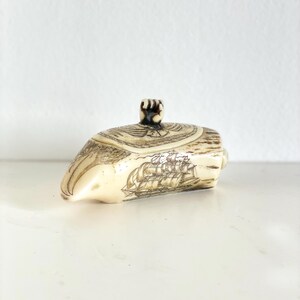 Op de afbeelding: Een crèmekleurige, gesneden container met een deksel. De container heeft de vorm van een boot, met gedetailleerde tekeningen van een zeilschip en andere nautische elementen. Het deksel heeft een kleine, donkere knop. Het is waarschijnlijk een decoratieve doos of een sieradenhouder.