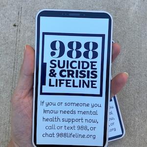 以下が含まれることがあります： スマートフォン型の青い縁取りのある白いステッカーで、「988 SUICIDE & CRISIS LIFELINE」というテキストと、「あなたまたはあなたが知っている人が今すぐメンタルヘルスのサポートを必要とする場合、988に電話またはテキストメッセージを送信するか、988lifeline.orgでチャットしてください。」というメッセージが書かれています。