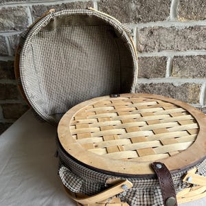 May include: Open picnic basket with a woven lid and a brown leather strap closure. The basket is made of light-colored wood and features a checkered fabric lining and trim. The lid is partially open, revealing the interior.