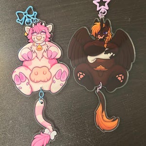 May include: Two acrylic charms featuring anthropomorphic characters. The left charm is a pink and white cow-like creature with a blue bow clasp. The right charm is a dark brown winged creature with orange hair and a purple star-shaped clasp.