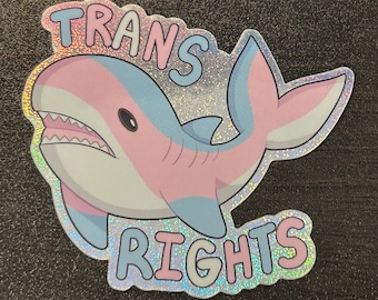 May include: A pink and blue shark with a glitter finish and the text "TRANS RIGHTS" in pink and blue letters.