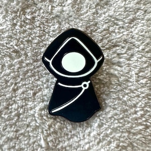 May include: Black and white enamel pin featuring a hooded figure with a white face and a black robe. The pin has a white outline.