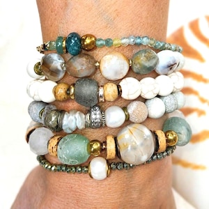 May include: A stack of five beaded bracelets featuring a variety of round, faceted, and irregular beads in shades of white, gray, tan, gold, and seafoam green. The bracelets include smooth stones, textured metallic accents, and clear crystal beads.