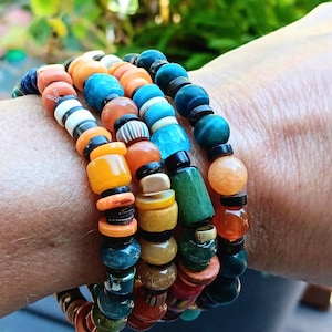 以下が含まれることがあります： オレンジ、青、緑、黒など、さまざまな色のビーズブレスレットのコレクション。 ブレスレットは、丸いビーズと円筒形のビーズが混在しており、重ね着したような外観になっています。 ブレスレットは手首に着用されています。