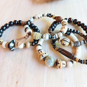 May include: Several beaded bracelets with a variety of designs. The bracelets incorporate black, white, and tan beads, with some featuring decorative elements such as pearls and star-shaped details. They are displayed on a light wooden surface.