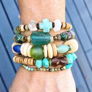 May include: A stack of five beaded bracelets featuring a variety of materials including a light blue cross-shaped bead, clear faceted beads, smooth green glass beads, tan wooden discs, dark brown faceted beads, and small turquoise-colored stones.