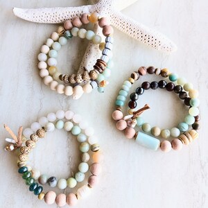 May include: Three layered beaded bracelets with a mix of light brown, beige, green, and blue beads. The bracelets are arranged on a white surface with a white starfish.