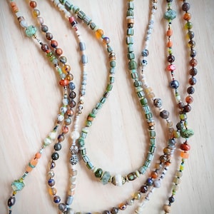 Peut inclure: Plusieurs colliers de perles de différentes couleurs, dont le vert, l'orange, le marron et le blanc, sont présentés sur un fond en bois clair. Les colliers présentent une variété de formes et de tailles de perles, créant un effet superposé. Les perles semblent être en verre et en pierre.