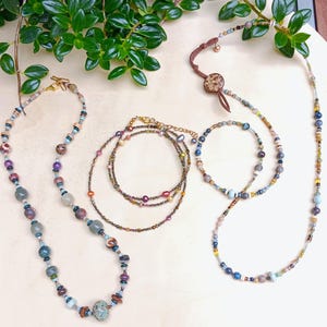 Pode incluir: Três colares com designs de contas variados. Um colar tem contas de pedra grandes e multicoloridas. Outro é um fio delicado de pequenas pérolas multicoloridas e contas metálicas. O terceiro é um cordão longo e fino com contas dispersas.