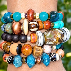 May include: A multi-strand bracelet featuring a diverse collection of beads in various shapes, sizes, and textures. The beads include polished turquoise, matte black, metallic copper, striped ceramic, and marbled glass elements with brass spacers.