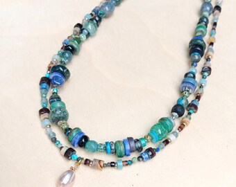 Peut inclure: Un collier multi-rangs composé d'un assortiment de perles dans des tons de bleu, vert, marron et gris. Le collier comprend diverses formes et textures de perles en verre et en pierre, terminé par un pendentif en perle rose pâle en forme de goutte.