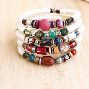 May include: A multi-strand bracelet featuring white disc-shaped beads interspersed with a variety of colorful accent beads. The accent beads include shades of red, blue, green, brown, and pink in various shapes, textures, and metallic finishes.